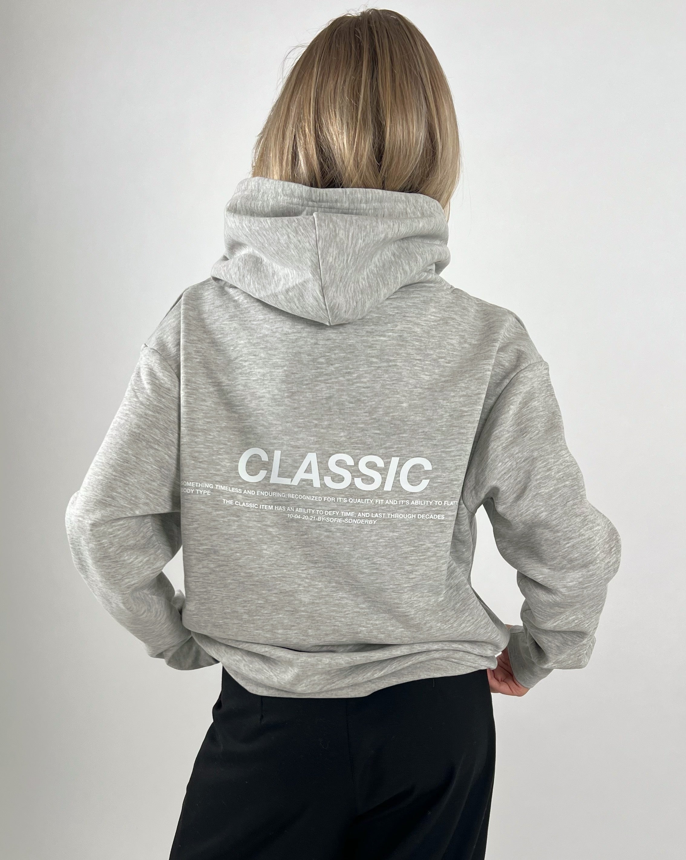En person iført en grå hættetrøje med teksten "CLASSIC" står med ryggen til i en neutral, lys baggrund. Mere tekst: "SOMETHING TIMELESS AND ENDURING, RECOGNIZED FOR ITS QUALITY, FIT, AND ITS ABILITY TO…"