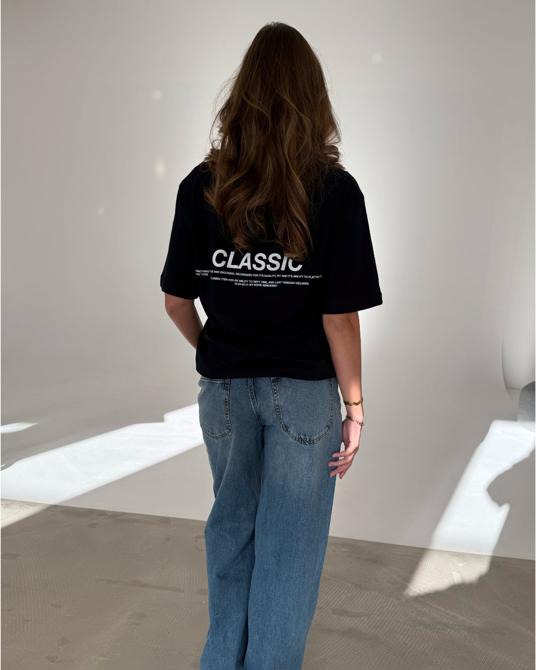 En person står med ryggen til iført en sort t-shirt med teksten "CLASSIC" og blå jeans i et oplyst rum uden dekoration.
