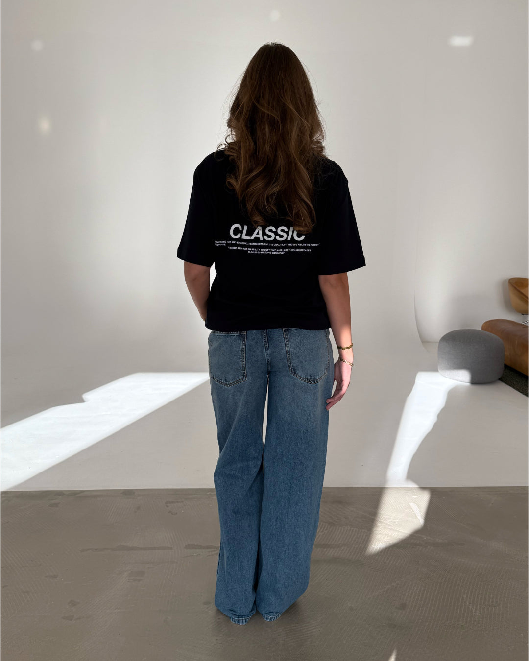 En person med langt hår og blå jeans står stille med ryggen til, iført en sort T-shirt med teksten "CLASSIC" i et minimalistisk, lyst rum.