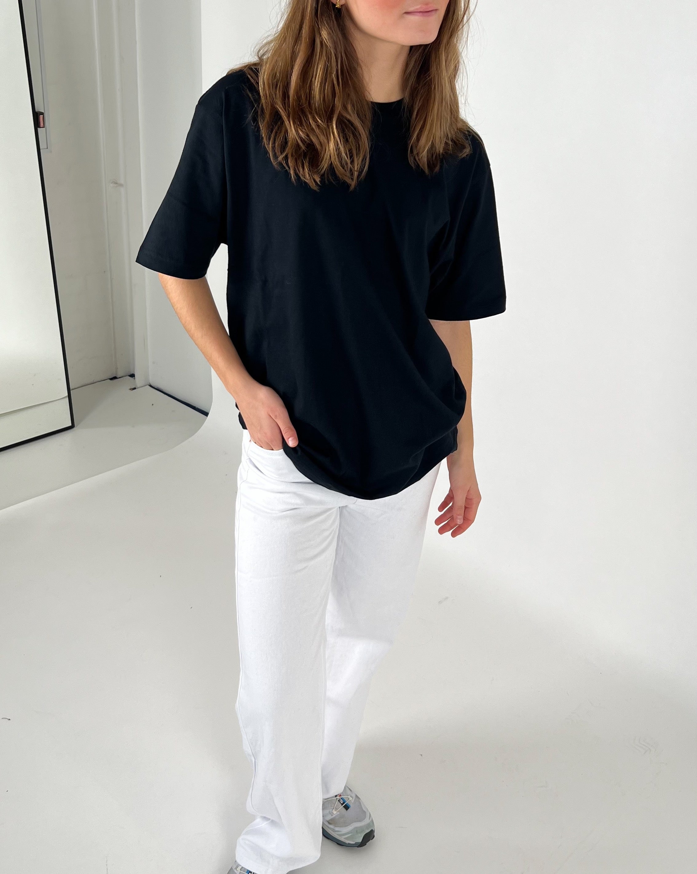 En person iført sort t-shirt og hvide bukser står afslappet i et minimalistisk rum med lyse vægge og gulv. Personen har en hånd i lommen og sneaker-sko på.