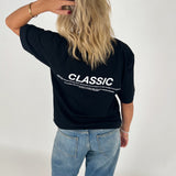 Kvinde i sort T-shirt med teksten "CLASSIC" står med ryggen til, rører sit hår. Hun bærer lyse jeans i en neutral, lys baggrund. Teksten på T-shirten er delvist læsbar.