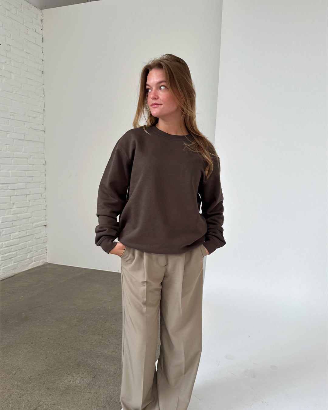 Kvinde iført brun sweatshirt og beige bukser står afslappet med hænderne i lommerne i et rum med hvide murstensvægge og gråt gulv.