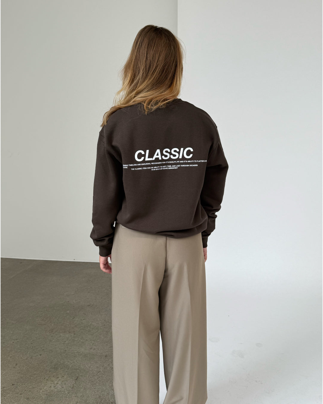 En person står med ryggen til iført en mørk trøje med teksten "CLASSIC" på ryggen. Personen befinder sig i et minimalistisk rum med hvid væg og gråt gulv.