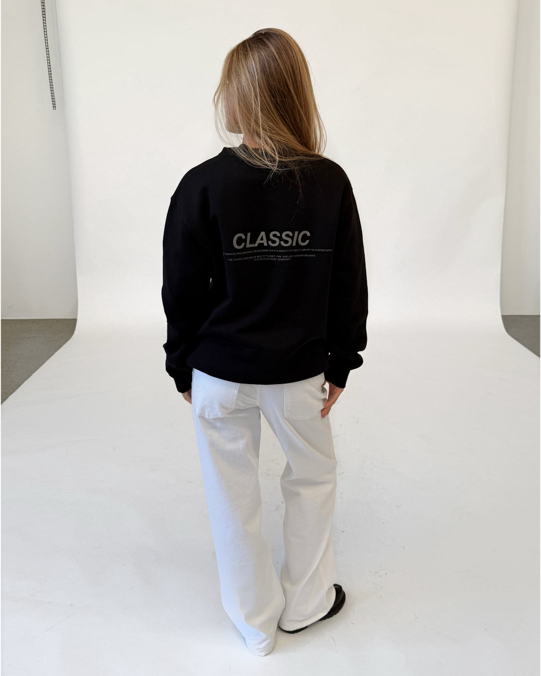 Modelfoto af pige i sort CLASSIC sweatshirt med reflekstryk, samt hvide NANNA jeans. Alt er fra BySofieSønderby.