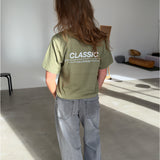 En person med langt brunt hår står i et lyst rum iført en grøn t-shirt med teksten "CLASSIC" på ryggen og grå jeans. Rummet har minimalistisk møblering.