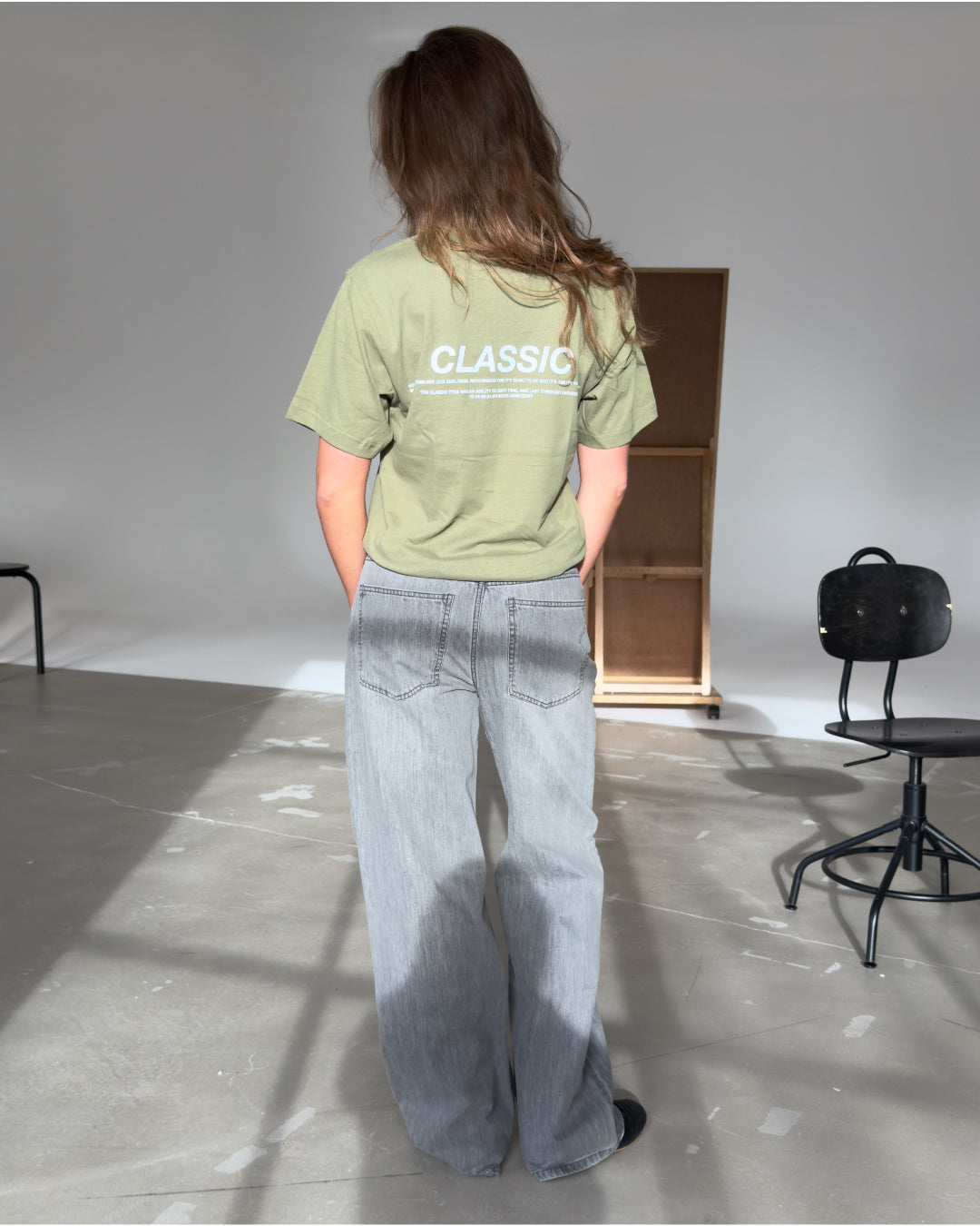 Person står med ryggen til iført en grøn T-shirt med teksten "CLASSIC" og lys-grå jeans, i et lyst rum med stole og en skillevæg i baggrunden.