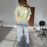 Person står med ryggen til iført en grøn T-shirt med teksten "CLASSIC" og lys-grå jeans, i et lyst rum med stole og en skillevæg i baggrunden.