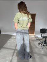 Person står med ryggen til iført en grøn T-shirt med teksten "CLASSIC" og lys-grå jeans, i et lyst rum med stole og en skillevæg i baggrunden.
