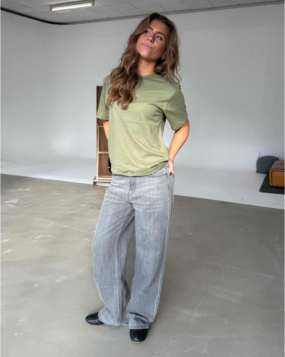 En person med langt, bølget hår bærer en grøn t-shirt og grå jeans i et lyst rum med en træhylde og mørkt møbel i baggrunden.