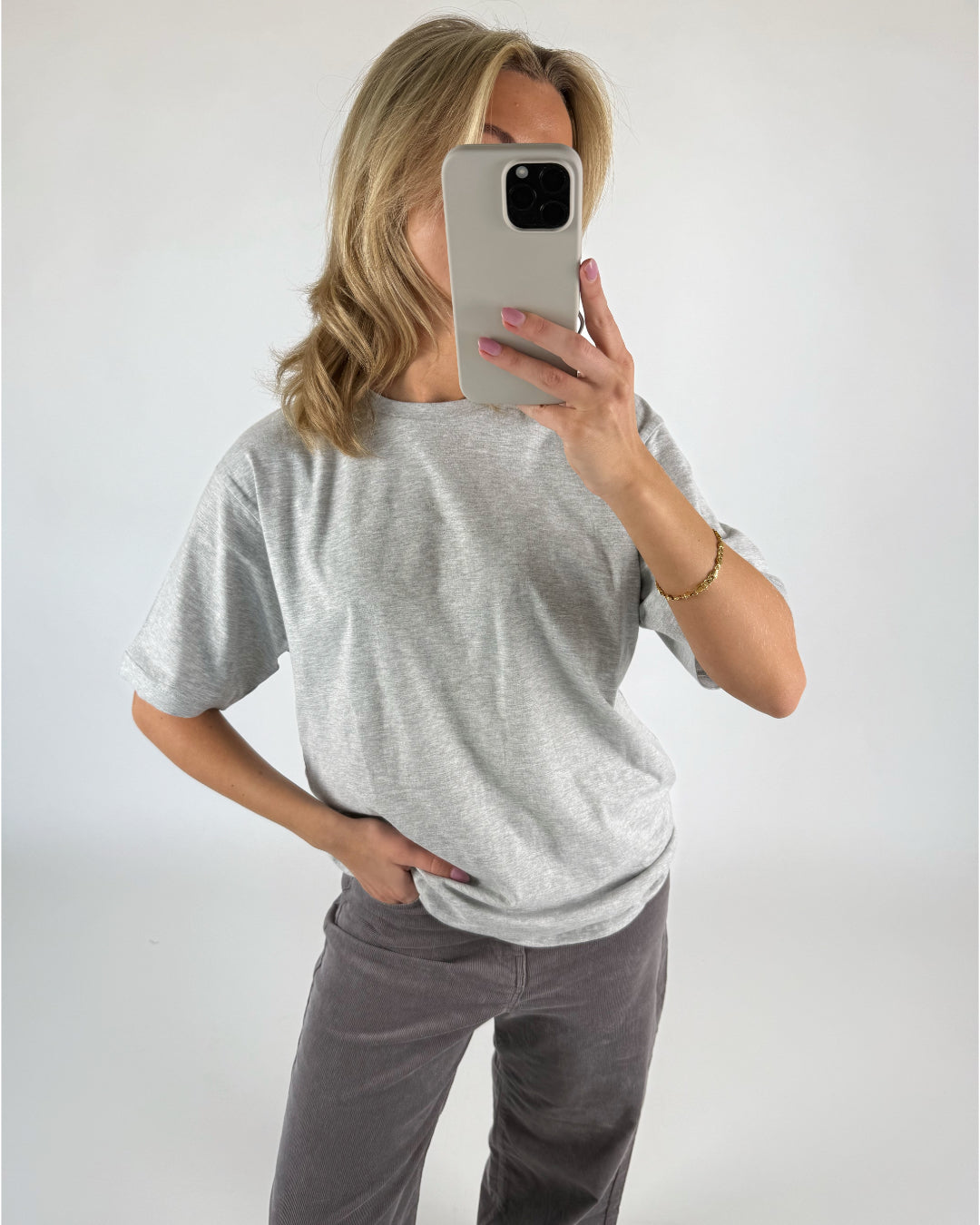 En person iført grå T-shirt og mørke bukser holder en smartphone foran ansigtet. Personen står i et lyst rum og ser ud til at tage et spejlbillede.