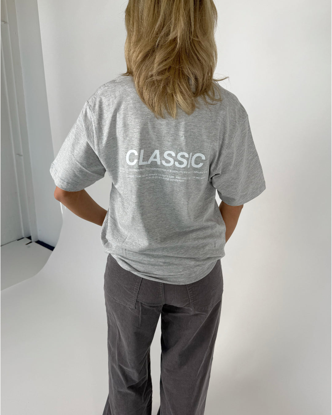 En person iført en grå T-shirt og mørke bukser står med ryggen til. På T-shirten står teksten "CLASSIC". Baggrunden er lys og enkel.