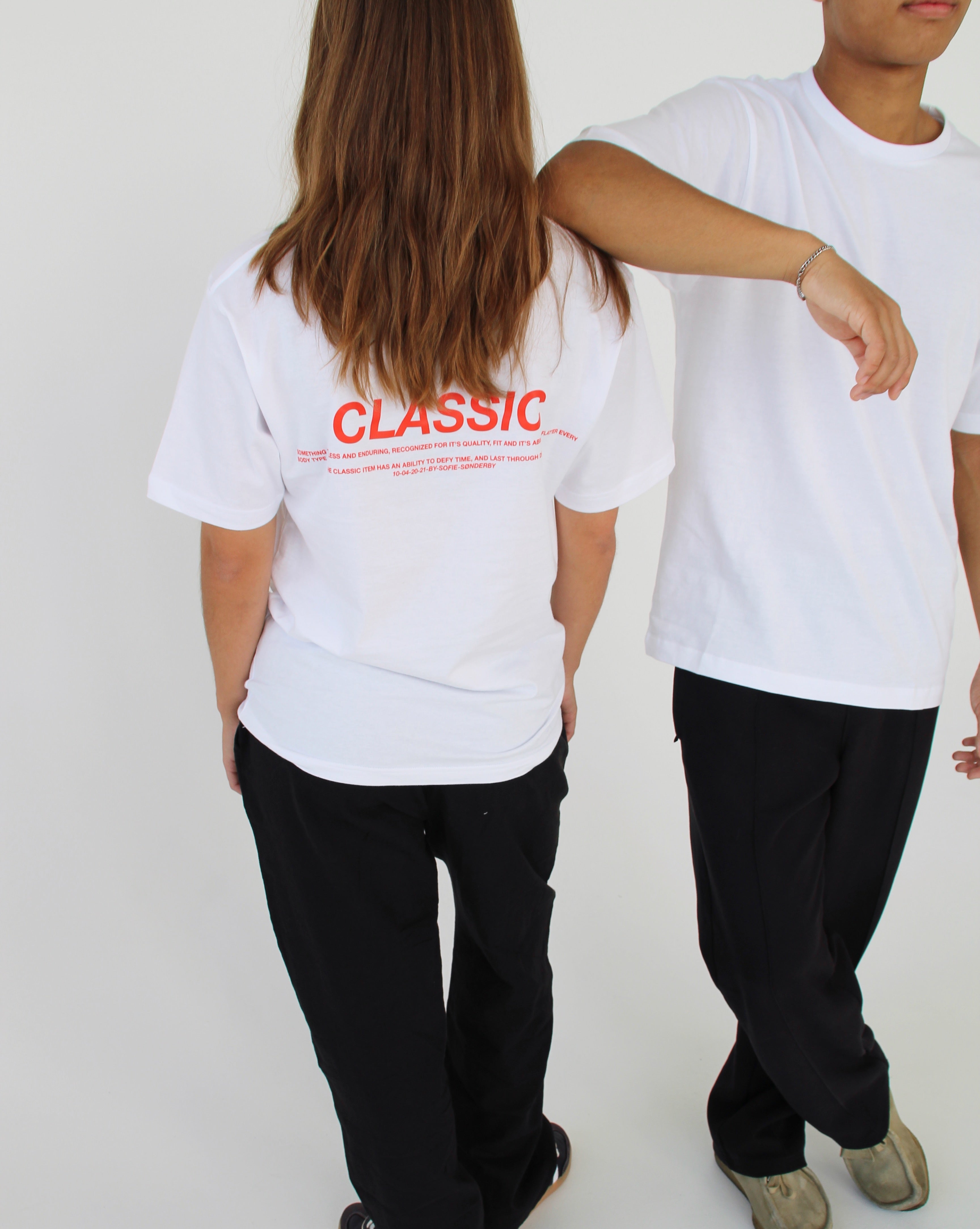 To personer iført hvide T-shirts og sorte bukser står mod en lys baggrund. Den ene T-shirt har tekst på ryggen: "CLASSIC" efterfulgt af yderligere ord i mindre skrift.