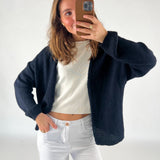 En person tager et selfie med en smartphone, iført en sort cardigan over en hvid bluse, og hvide bukser. Personen står i et lyst indendørs rum.