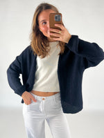 En person tager et selfie med en smartphone, iført en sort cardigan over en hvid bluse, og hvide bukser. Personen står i et lyst indendørs rum.