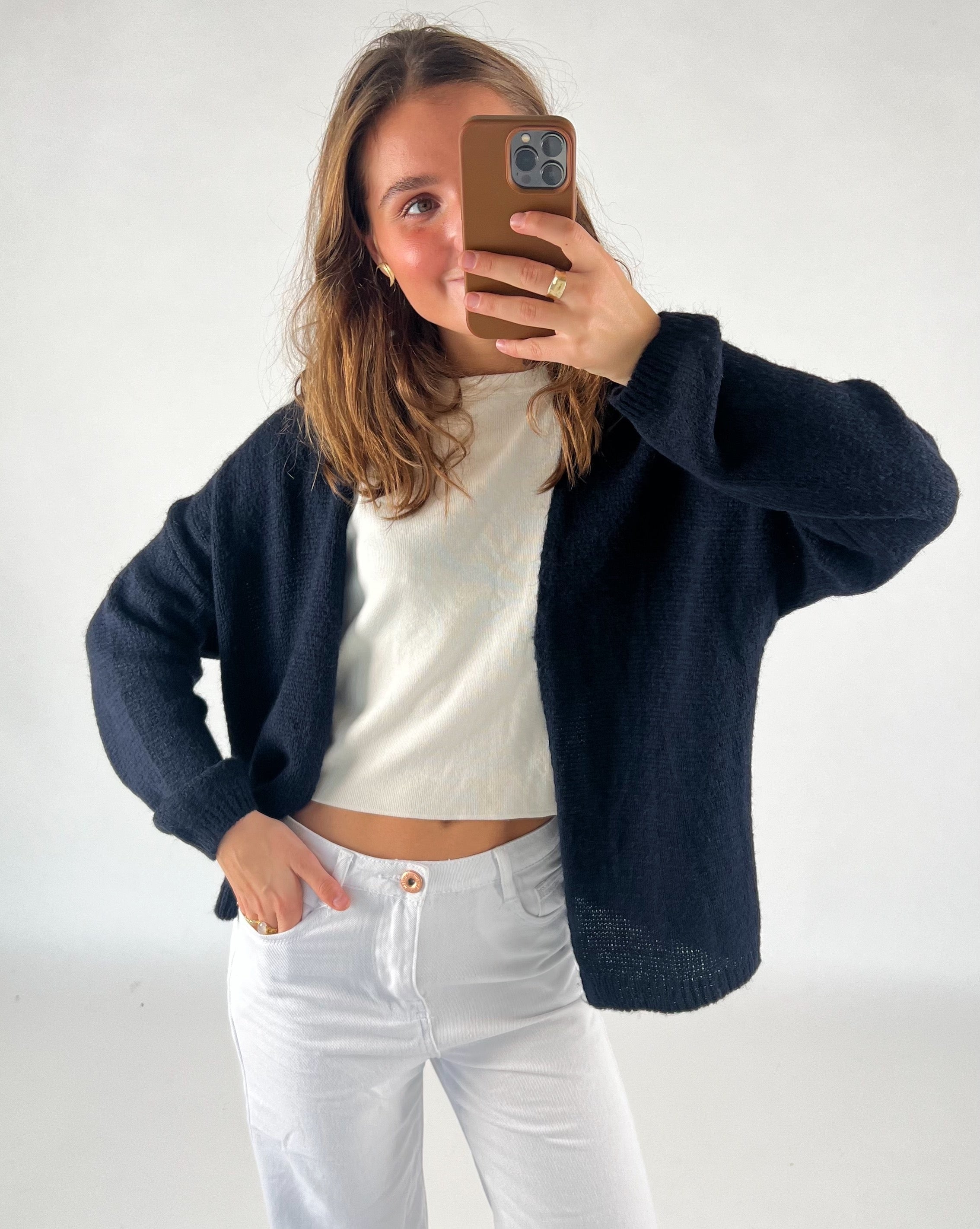En person tager et selfie med en smartphone, iført en sort cardigan over en hvid bluse, og hvide bukser. Personen står i et lyst indendørs rum.