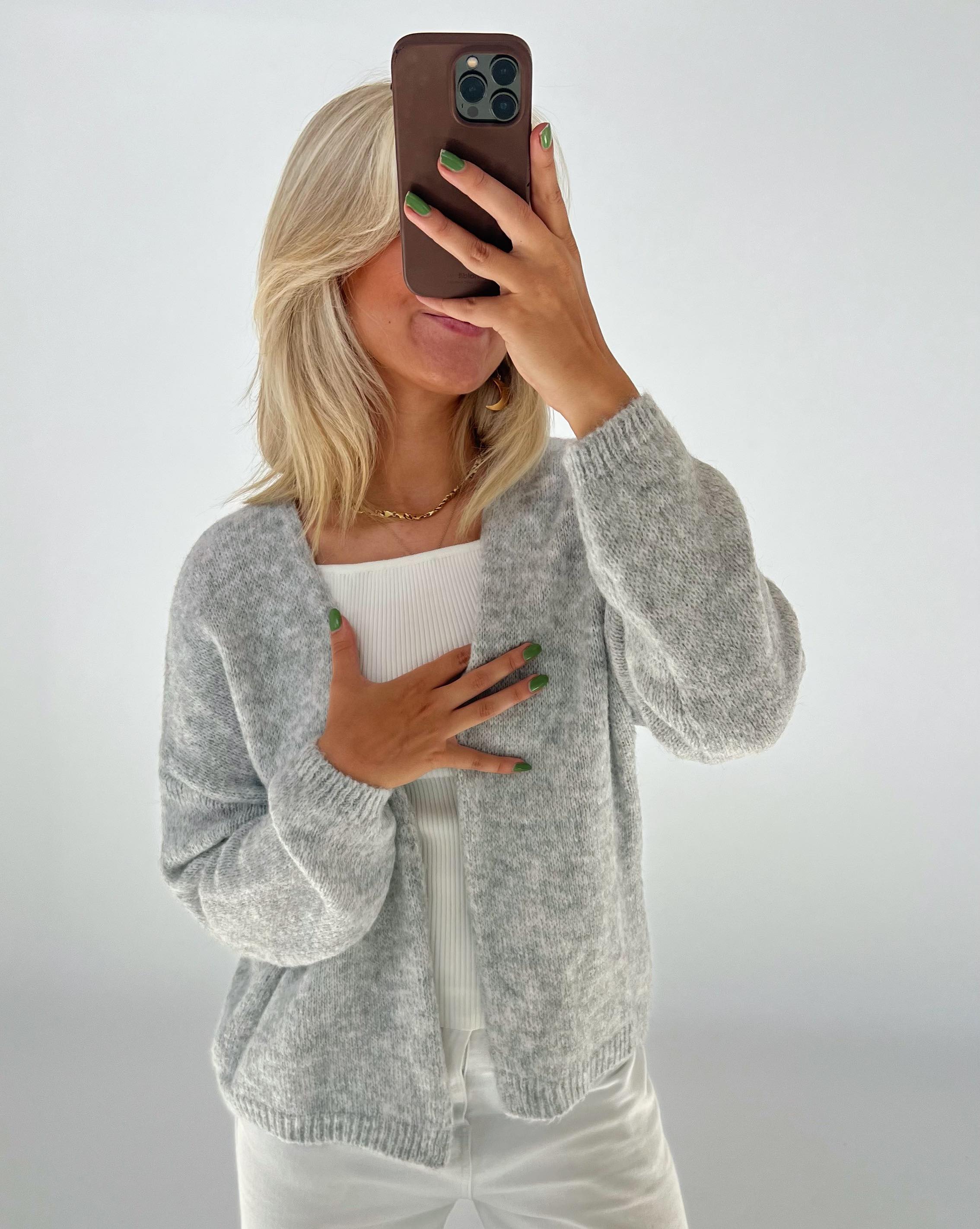 Person tager selfie; iført grå cardigan; holder mobil til ansigt; lyse lokker, grøn neglelak; hvide bukser; hvid baggrund.