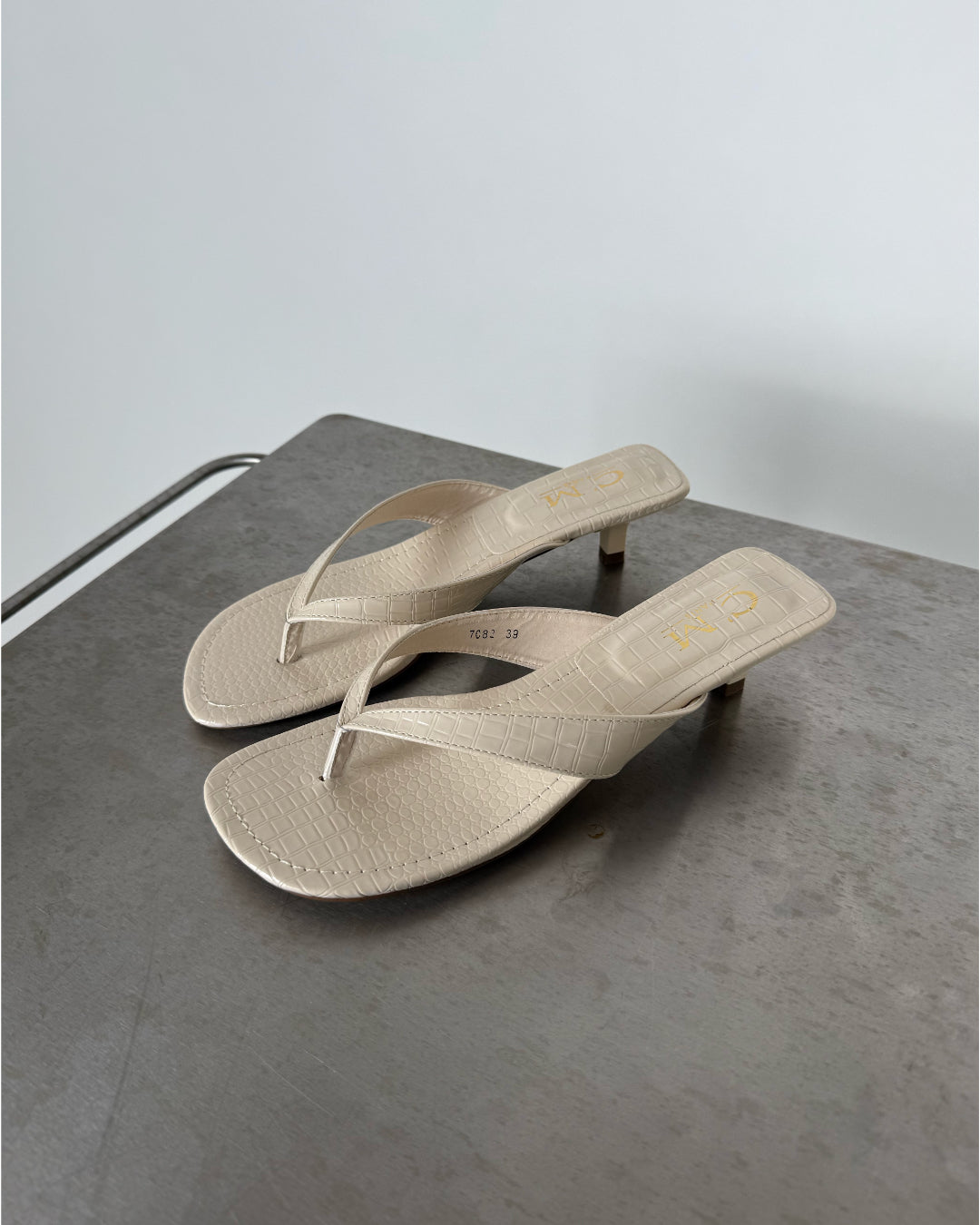 Beige sandaler med hæl står på en grå metaloverflade. Venstre sandal har tekst "7C82 39", højre "CII". Baggrunden er enkel og lysegrå.