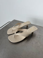 Beige sandaler med hæl står på en grå metaloverflade. Venstre sandal har tekst "7C82 39", højre "CII". Baggrunden er enkel og lysegrå.