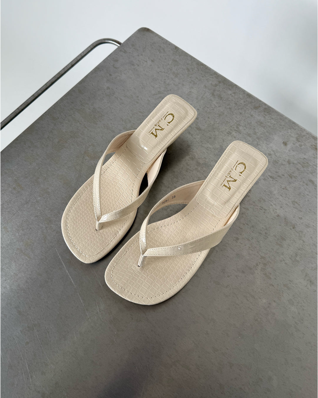 Beige sandaler med krokodillemønster ligger på en grå overflade. Tekst på indersålen viser "CM Made in China".