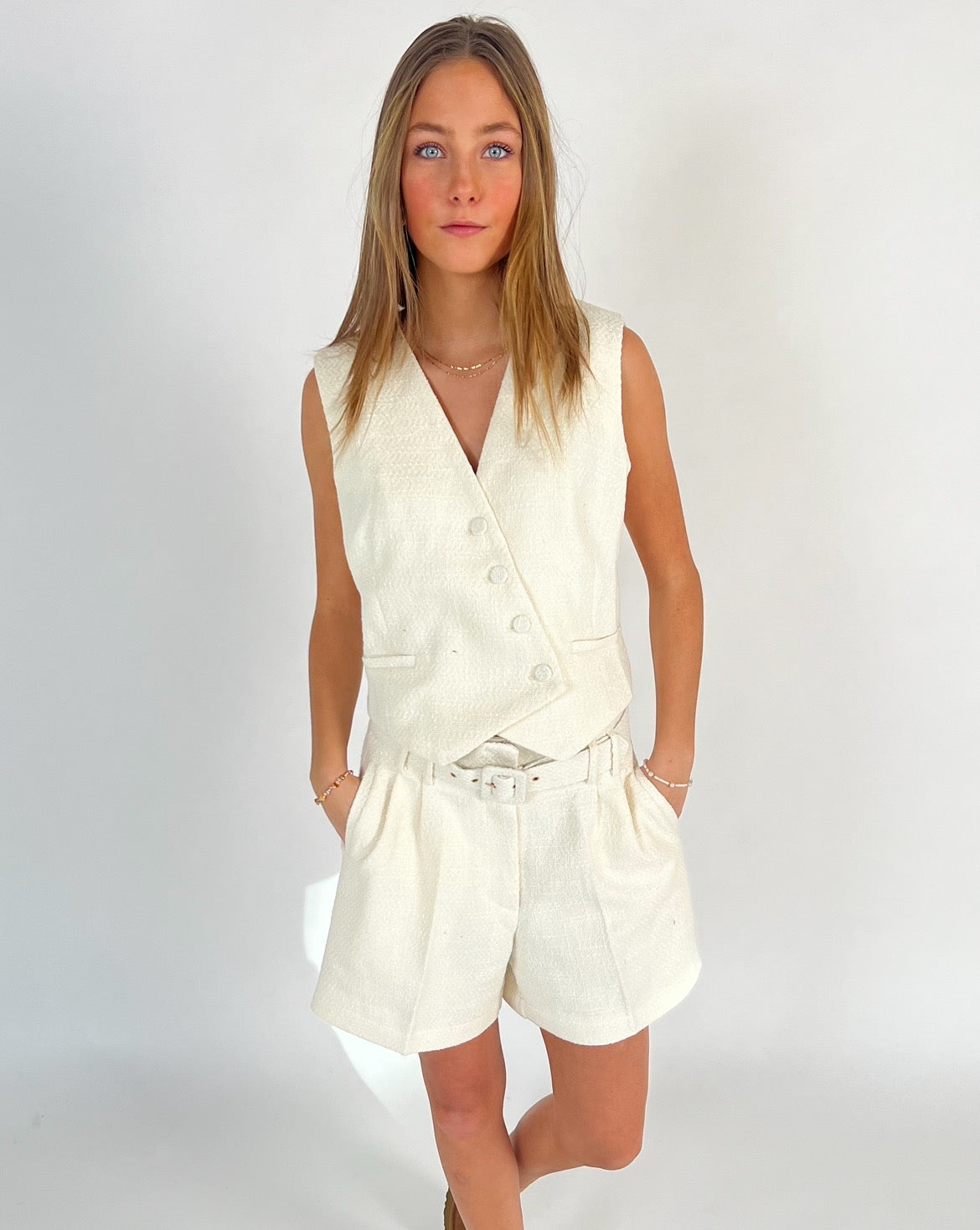 En person bærer et hvidt ærmeløst vest-og-shorts sæt og står i et minimalistisk hvidt rum. Hænderne er i lommerne, og personen ser direkte fremad.