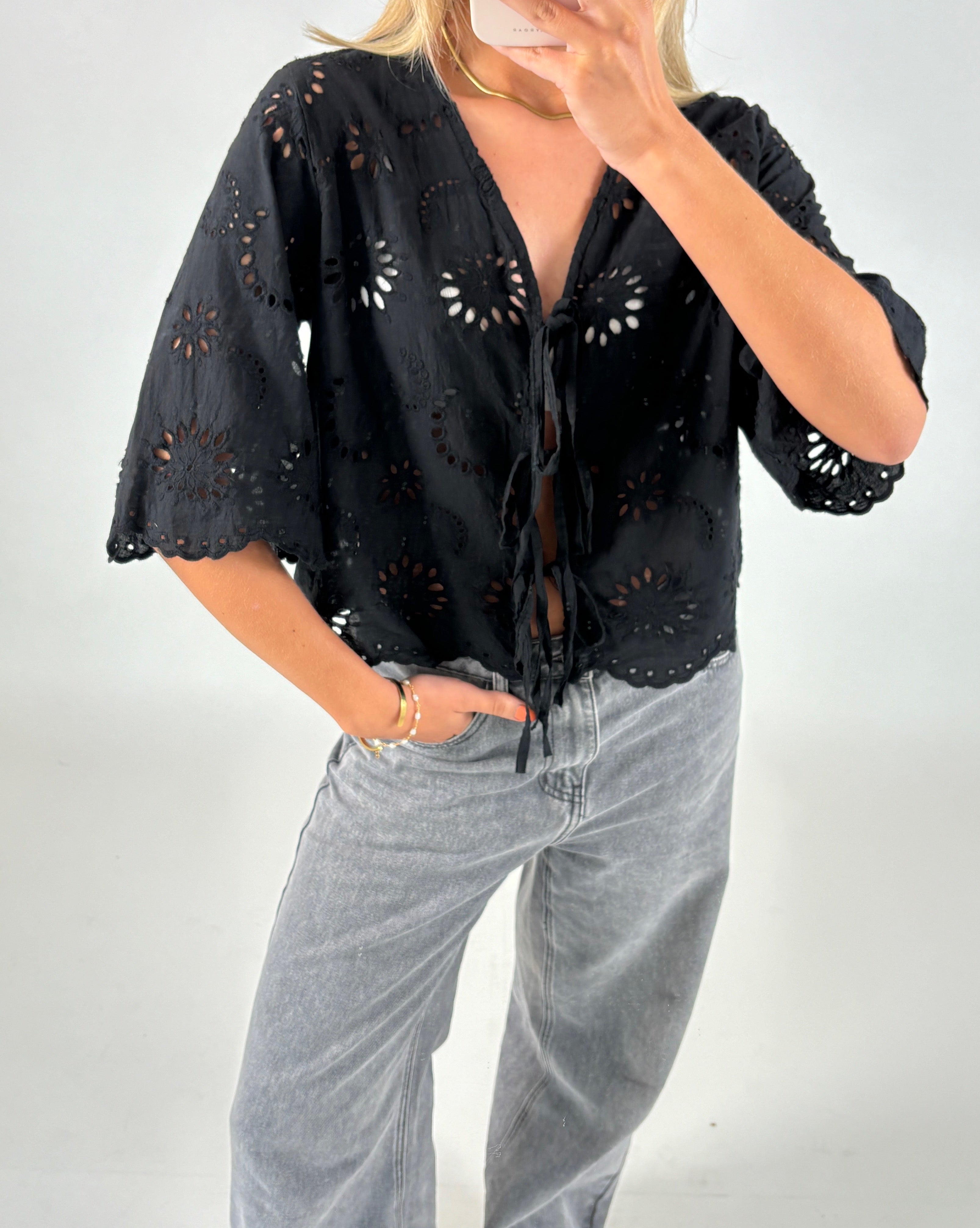En person iført en sort bluse med blomsterbroderi står med hånden i lommen på grå jeans, poserende mod en lys baggrund.
