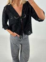 Kvinde iført sort bluse med broderede blomstermønstre, står med en hånd i lommen på grå jeans. Baggrunden er neutral, og hendes hår er blondt og skulderlangt.