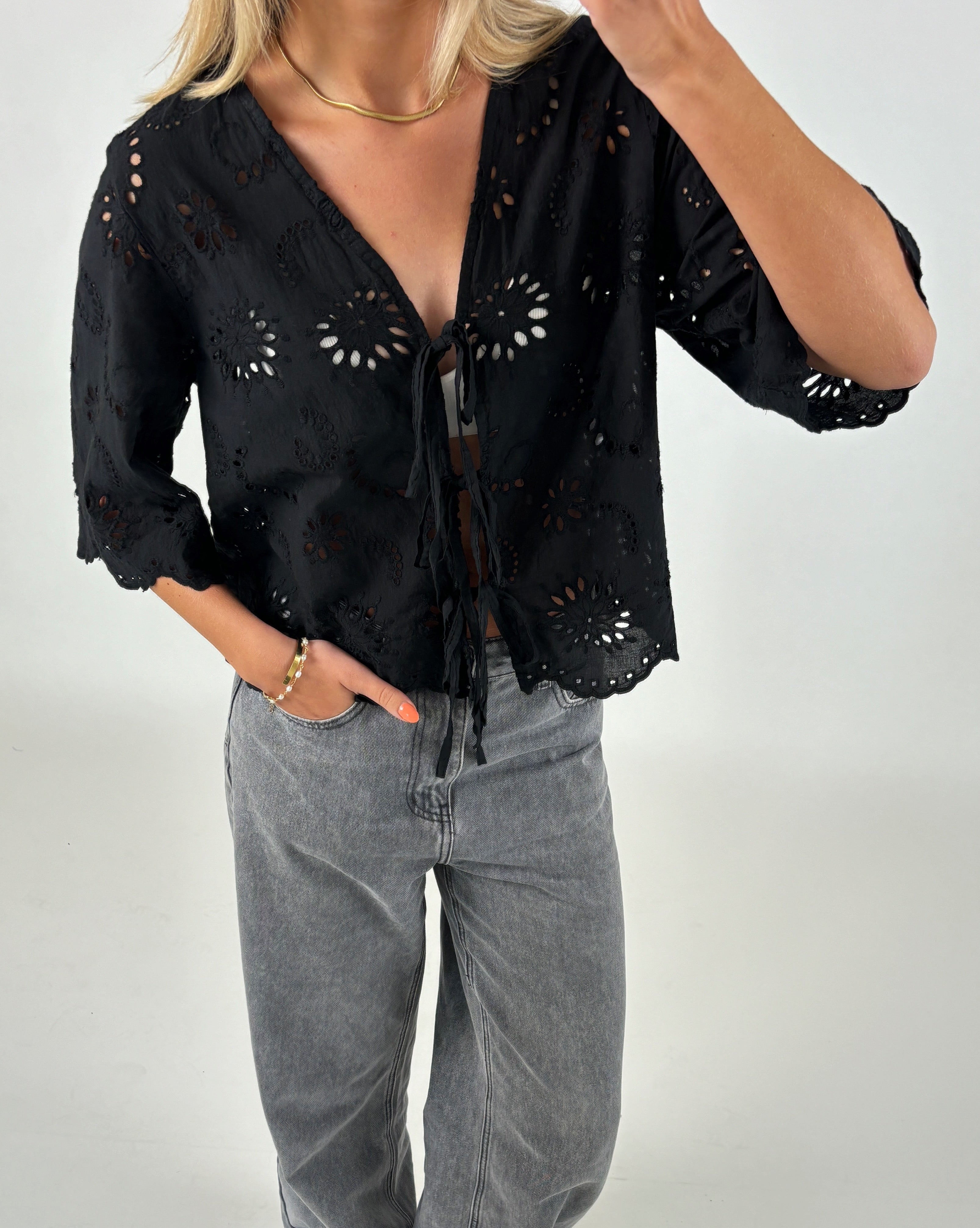 Kvinde iført sort bluse med broderede blomstermønstre, står med en hånd i lommen på grå jeans. Baggrunden er neutral, og hendes hår er blondt og skulderlangt.