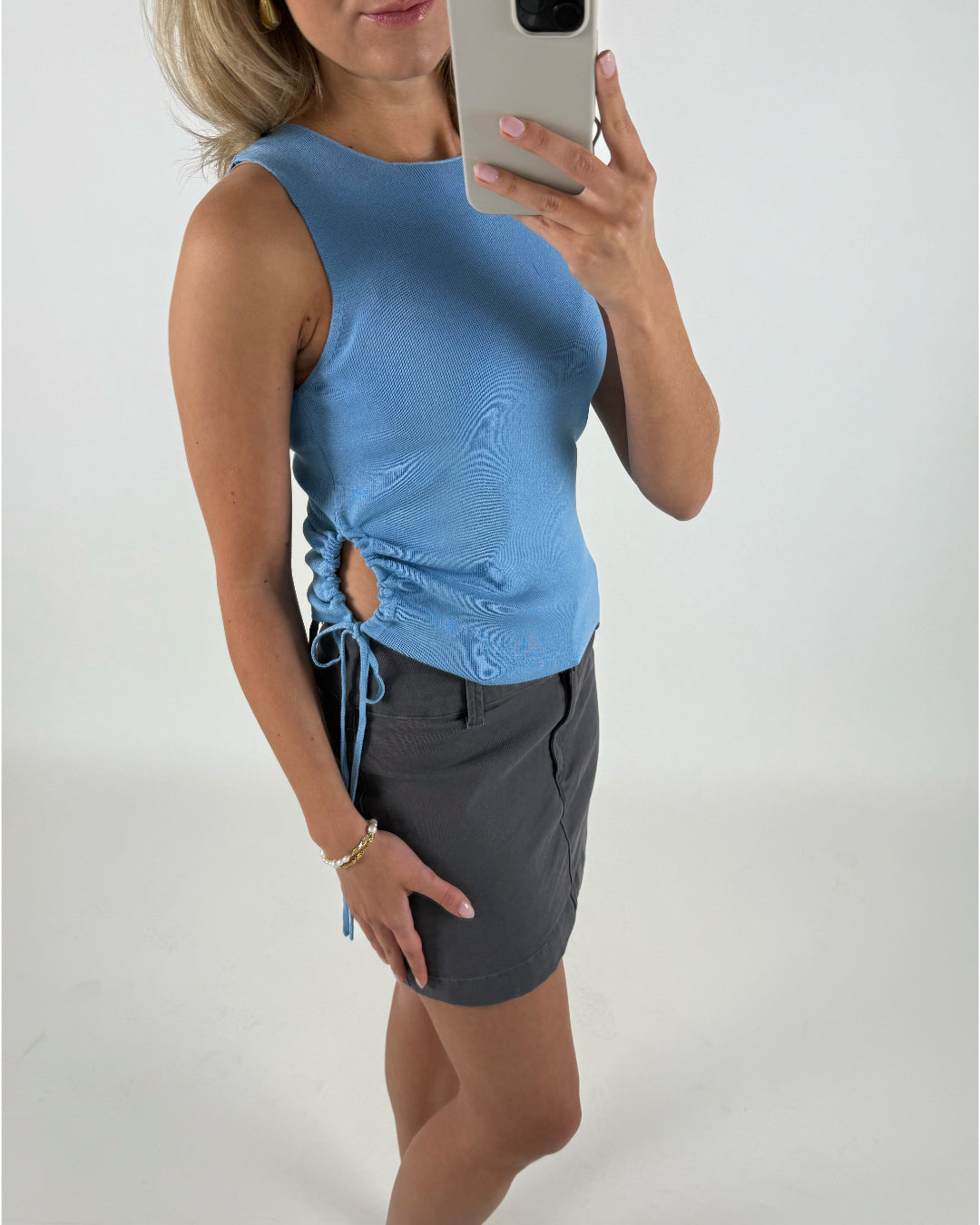 En person i en lyseblå top og mørk nederdel tager en selfie med en smartphone. Personen bærer armbånd, og baggrunden er en neutral, lys flade.