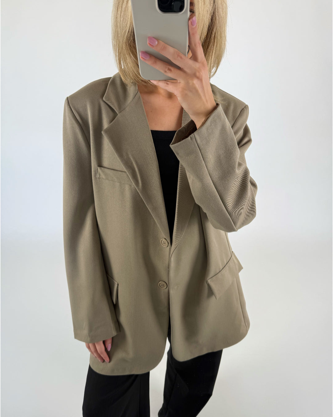 En person iført en beige blazer holder en smartphone op foran ansigtet. Personen står i et rum med en neutral, hvid baggrund.