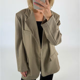 En person iført en beige blazer holder en smartphone op foran ansigtet. Personen står i et rum med en neutral, hvid baggrund.