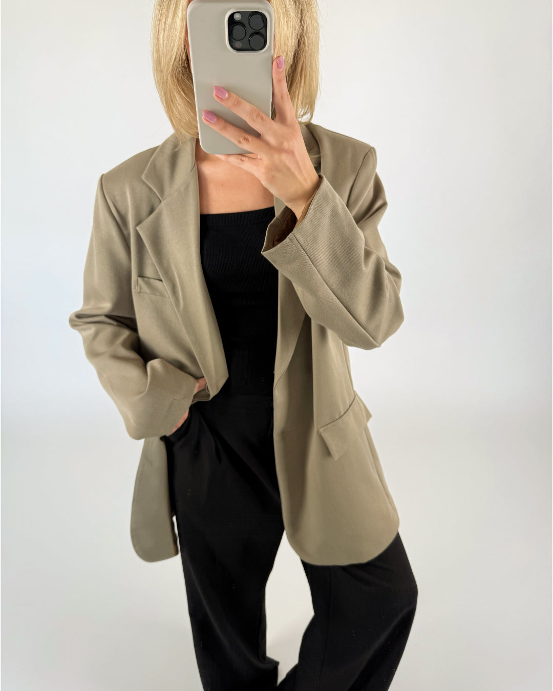 En person iført beige jakke og sort tøj tager et spejlfoto med en mobiltelefon, mens de står i et minimalistisk, hvidt rum.