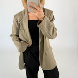 En person iført beige jakke og sort tøj tager et spejlfoto med en mobiltelefon, mens de står i et minimalistisk, hvidt rum.