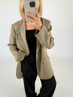 En person iført beige jakke og sort tøj tager et spejlfoto med en mobiltelefon, mens de står i et minimalistisk, hvidt rum.