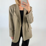 En person i beige blazer tager et spejlbillede med en smartphone, iført sort top og bukser; omgivelserne er et hvidt, enkelt rum.