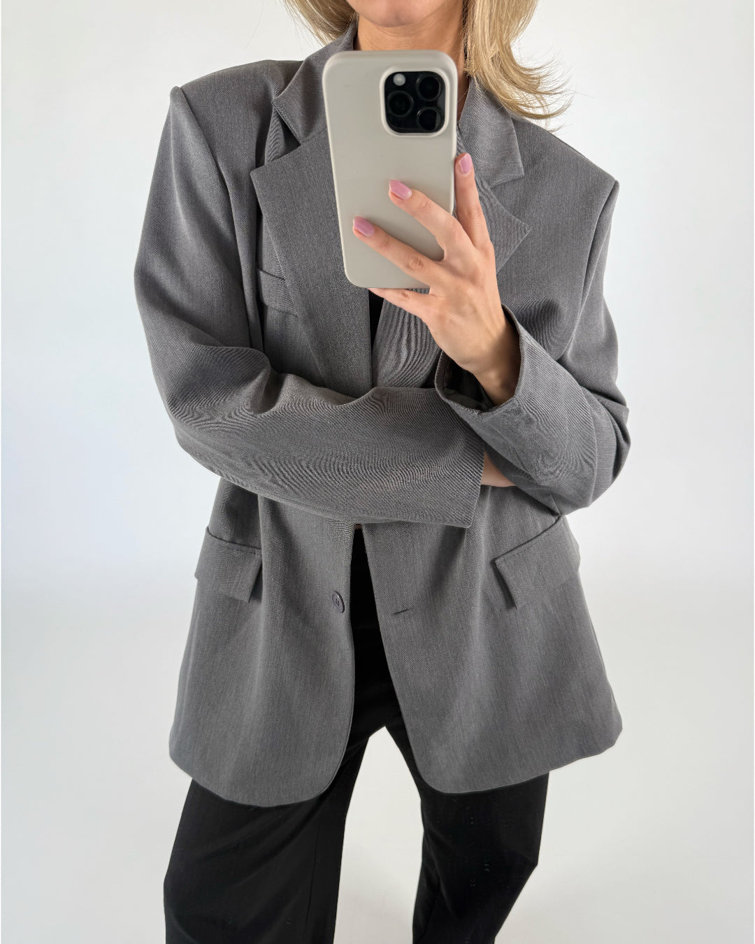 En person i en grå blazer tager et selfie med en smartphone. Personen står i et minimalistisk rum med en hvid baggrund.