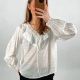 En person iført en hvid bluse med blondekant tager et spejlbillede med en smartphone i brun cover; baggrunden er neutral.