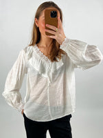 En person iført en hvid bluse med blondekant tager et spejlbillede med en smartphone i brun cover; baggrunden er neutral.