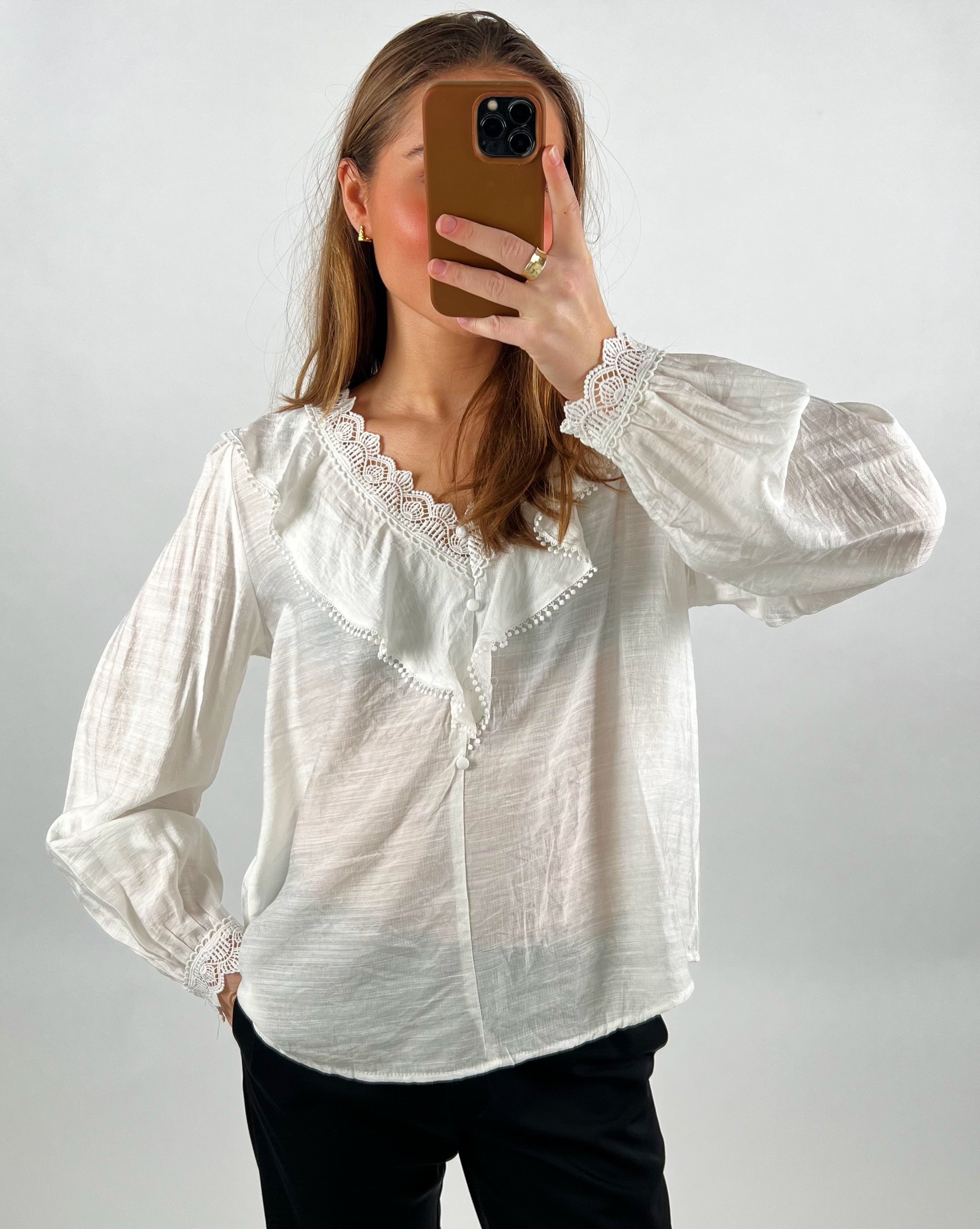 En person iført en hvid bluse med blondekant tager et spejlbillede med en smartphone i brun cover; baggrunden er neutral.