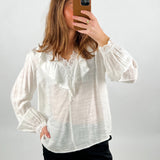 En person bærer en hvid bluse med blondekant, tager et spejlselfie med en smartphone i brun etui, stående mod en neutral baggrund.