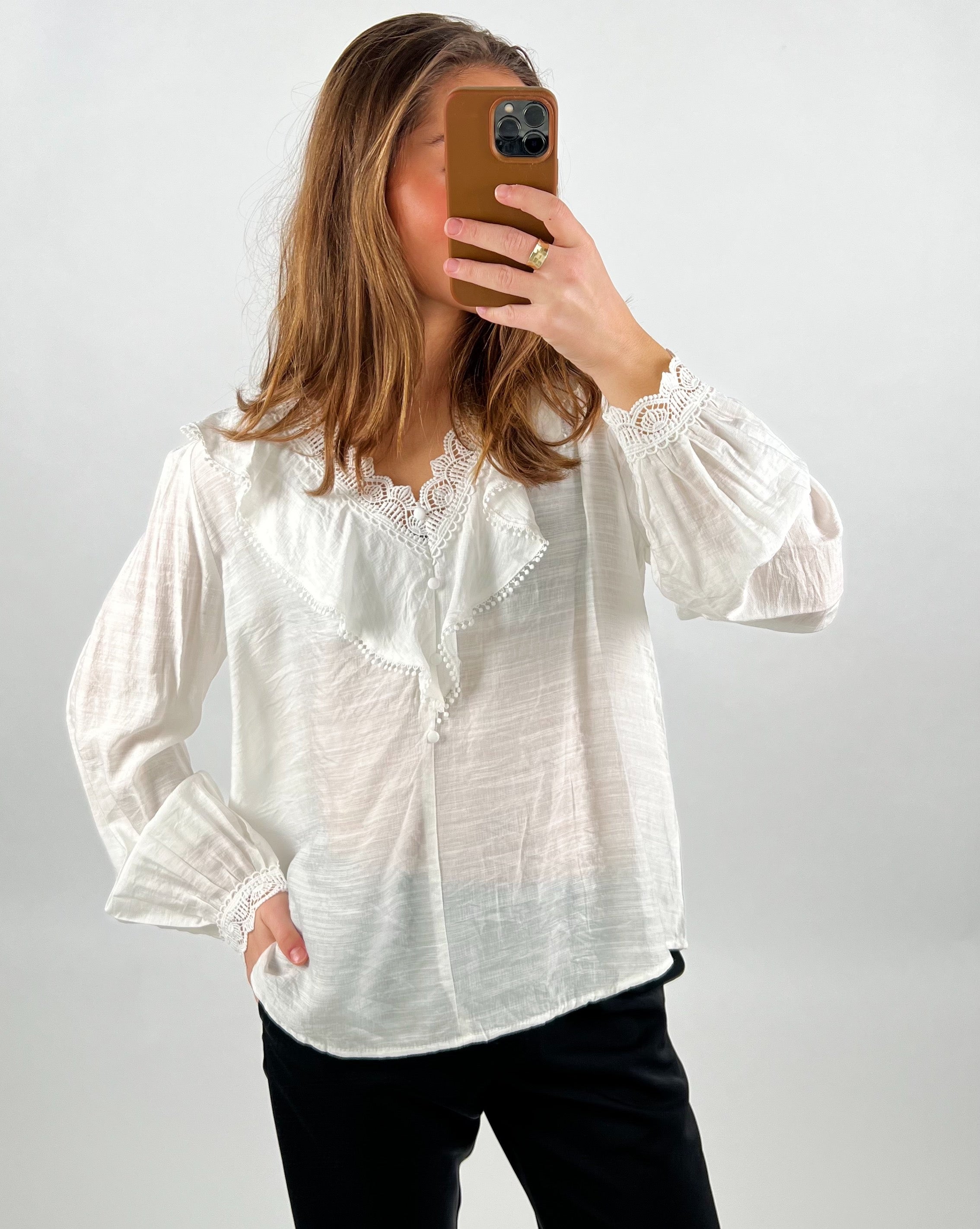 En person bærer en hvid bluse med blondekant, tager et spejlselfie med en smartphone i brun etui, stående mod en neutral baggrund.