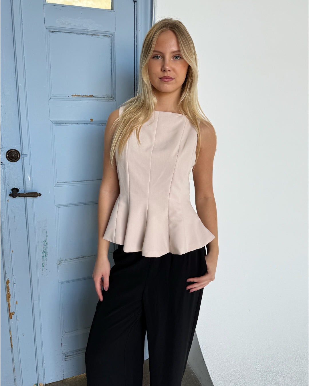 En kvinde står foran en lys blå dør iført en lys beige top og sorte bukser, med en neutral baggrund.
