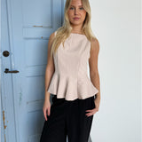En kvinde står foran en lys blå dør iført en lys beige top og sorte bukser, med en neutral baggrund.
