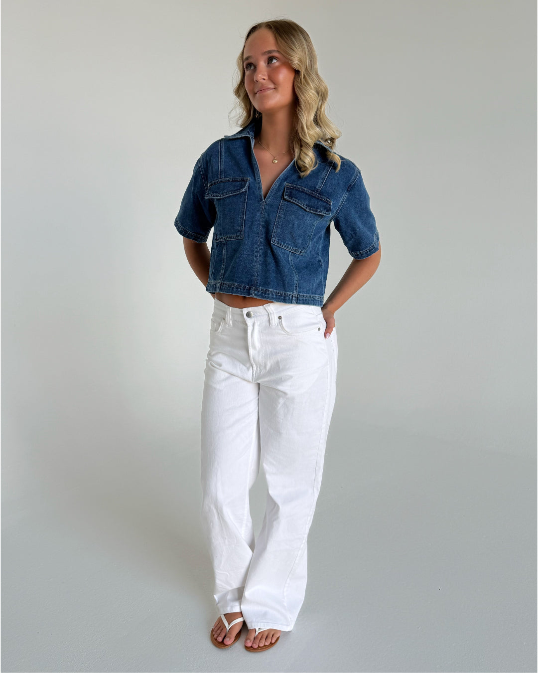 En person står afslappet iført en blå denim skjorte og hvide bukser, med hænderne i bukselommerne, i et minimalistisk, hvidt rum.