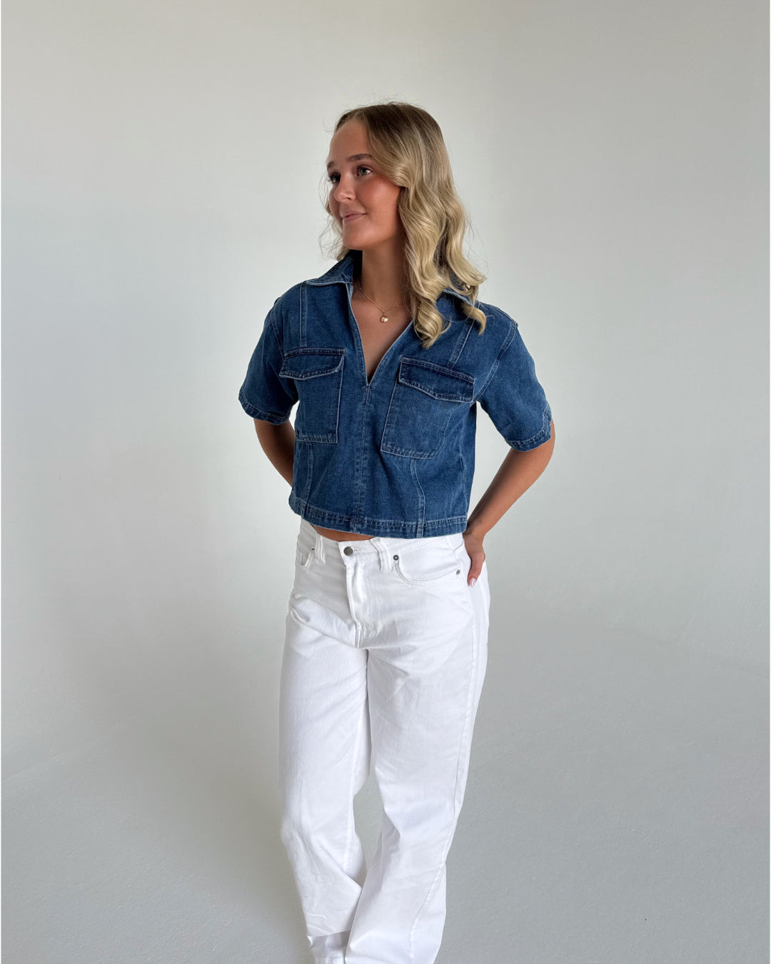 En kvinde iført en blå denimskjorte og hvide bukser står med hænderne bag ryggen i et neutralt, lyst rum. Hun kigger til venstre.