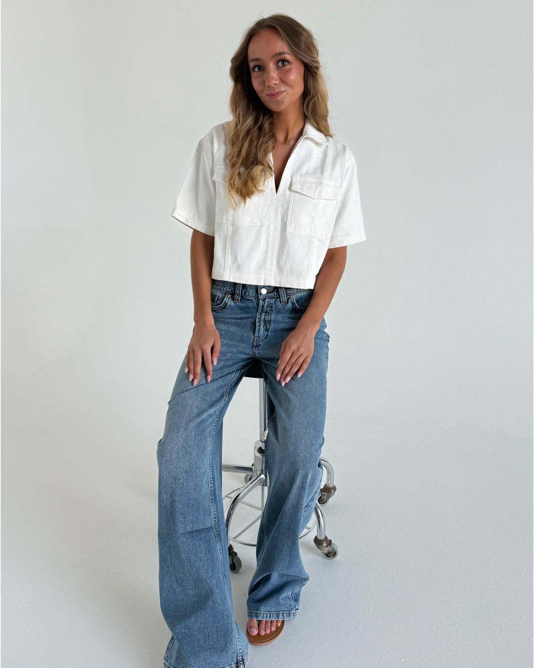 Kvinde sidder på en metalstol i et hvidt rum. Hun har en hvid crop-top og blå jeans på. Håret er langt og bølget, og hun smiler let.