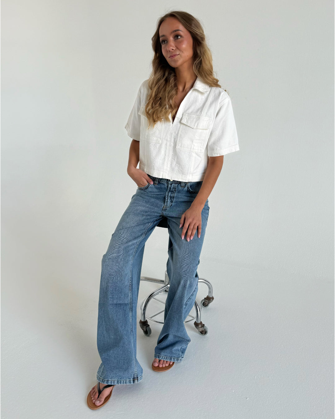 Kvinde står afslappet og hviler armen på en høj stol i et minimalistisk rum. Hun bærer en hvid kortærmet skjorte, blå jeans og sandaler. Ingen tekst til stede.