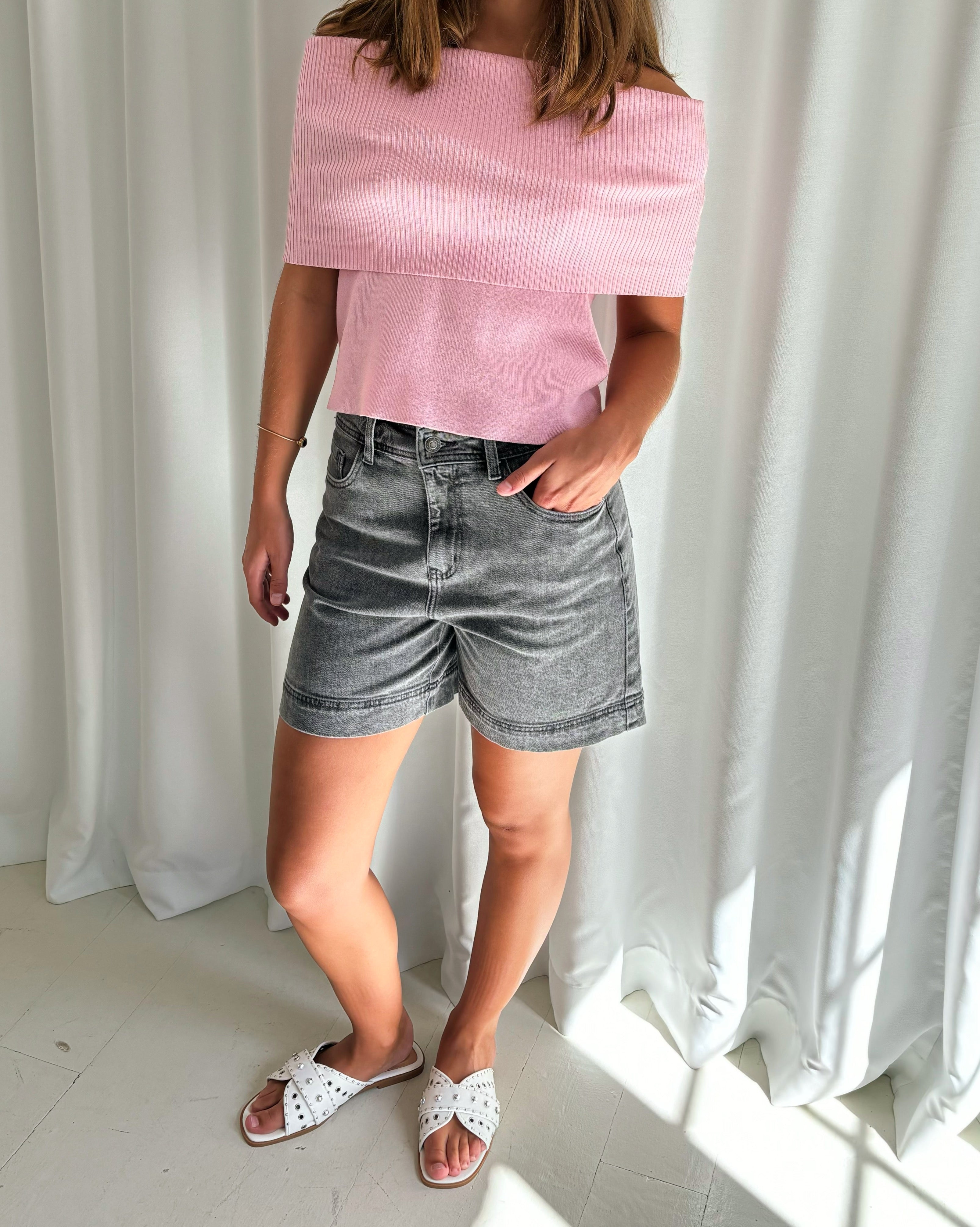 En person bærer en lyserød off-shoulder top og grå shorts, står på et lyst gulv foran en hvid gardinbaggrund. De har hvide, dekorerede sandaler på fødderne.
