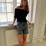 Ung kvinde står afslappet ved et vindue iført sort bluse og grå shorts, smiler i et lyst rum med radiator og rørinstallationer.