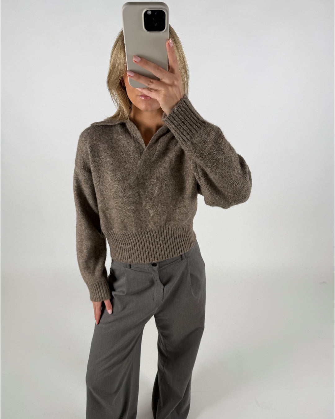 En person iført en brun sweater og grå bukser tager et selfie med en smartphone i et lyst rum.