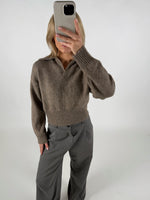 En person iført en brun sweater og grå bukser tager et selfie med en smartphone i et lyst rum.